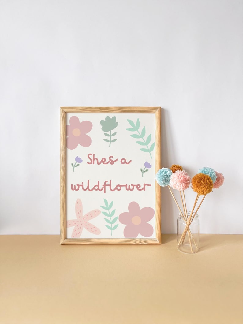 May include: A framed print with the text "She's a wildflower" and floral illustrations in pink, green, and purple. Beside it, a glass jar holds several pom-pom flowers in various pastel colors.
