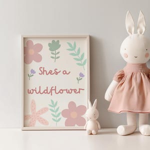 May include: A framed print with the text "Shes a wildflower" and floral illustrations in pink, green, and purple. A large stuffed bunny wearing a pink dress and a small bunny figurine are next to the print.