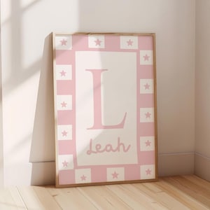 May include: A framed art print with a pink and white design. The print has a pink border with white stars and squares. The centre has a large pink letter "L" and the name "Leah" in a cursive font. The frame is a light wood colour.