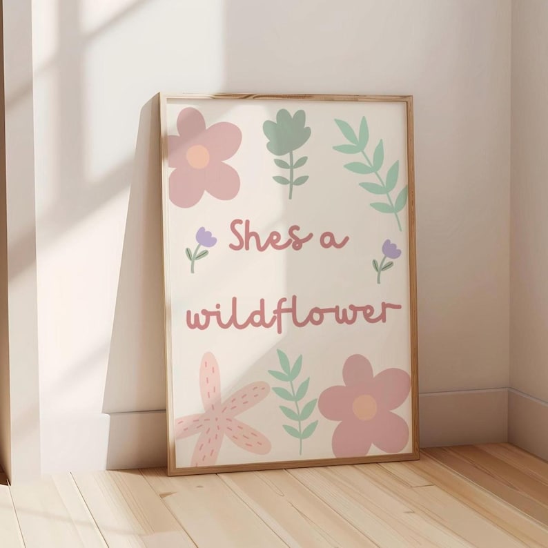 May include: A framed art print with the text "Shes a wildflower" in a cursive font. The print features pastel pink, purple, and green floral illustrations on a cream background. The frame is a light brown colour.