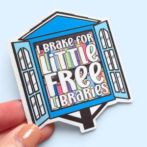 May include: A colorful sticker featuring a whimsical design of a little free library. The sticker has a blue roof and open windows with the text "I Brake for Little Free Libraries" in a playful font. The windows are filled with colorful book spines.
