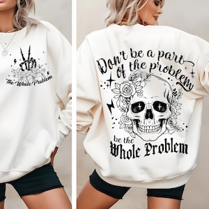 Peut inclure: Sweat-shirt crème avec des motifs graphiques noirs. L'avant présente une main de squelette faisant le signe de la paix avec des accents floraux et le texte "The Whole Problem". Le dos affiche un crâne avec des éléments floraux et le texte "Don't be a part of the problem, be the Whole Problem."
