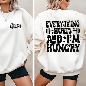 May include: White sweatshirt with a black dumbbell graphic on the front and the text "EVERYTHING HURTS AND I'M HUNGRY" on the back. The text is in a retro font with stars.