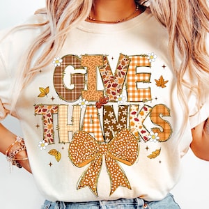 Peut inclure: Sweat-shirt crème avec l'inscription "GIVE THANKS" dans un design sur le thème de l'automne. Les lettres sont remplies de motifs à carreaux, floraux et léopard. Un grand nœud et des feuilles d'automne complètent le design.
