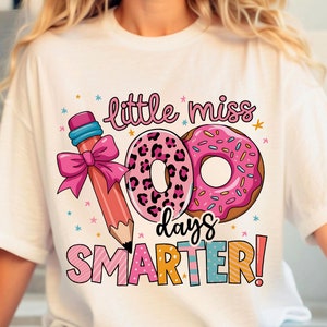 Puede incluir: Camiseta blanca con un gráfico rosa, amarillo, azul y naranja que dice "little miss 100 days smarter!" con un lápiz rosa, una rosquilla rosa con chispas y una rosquilla de leopardo rosa y negro.
