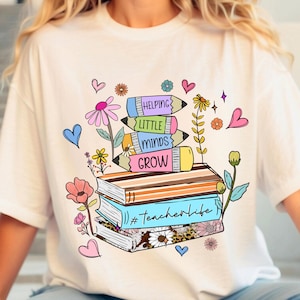 May include: White t-shirt with a colorful graphic of stacked books, pencils, and flowers. The pencils have the words "Helping Little Minds Grow" written on them. The bottom book has the hashtag "#teacherlife" written on it.