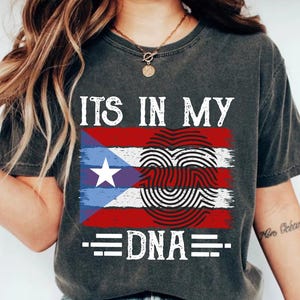 Puede incluir: Camiseta gris oscuro con un diseño de la bandera de Puerto Rico y el texto "ITS IN MY DNA". El diseño de la bandera incluye rayas rojas y blancas, un triángulo azul con una estrella blanca y un corazón de huellas dactilares.