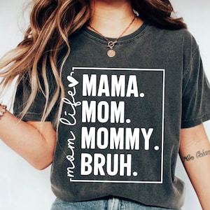 May include: A dark grey t-shirt with a white graphic that says "mama life" with a heart symbol, "mama", "mom", "mommy", and "bruh" in a white rectangle.