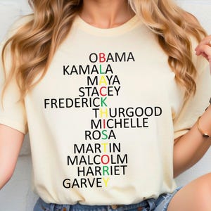 Może przedstawiać: Ż&oacute;łta koszulka z czarnym, zielonym i czerwonym wzorem, kt&oacute;ry tworzy napis "BLACK" z nazwiskami wybitnych Afroamerykan&oacute;w: Obama, Kamala, Maya, Stacey, Frederick, Thurgood, Michelle, Rosa, Martin, Malcolm, Harriet i Garvey.