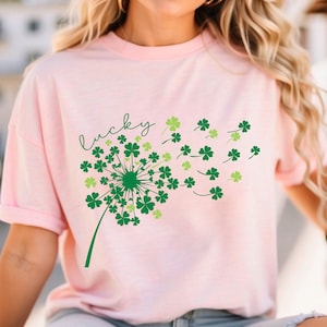 Puede incluir: Una camiseta rosa con un dise&ntilde;o de diente de le&oacute;n de tr&eacute;bol verde y la palabra "lucky" en letra cursiva.