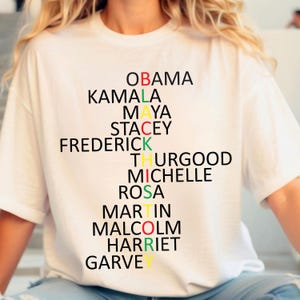 Może przedstawiać: Biała koszulka z czarno-kolorowym wzorem układanki słownej. Słowa to "OBAMA", "KAMALA", "MAYA", "STACEY", "FREDERICK", "THURGOOD", "MICHELLE", "ROSA", "MARTIN", "MALCOLM", "HARRIET" i "GARVEY".