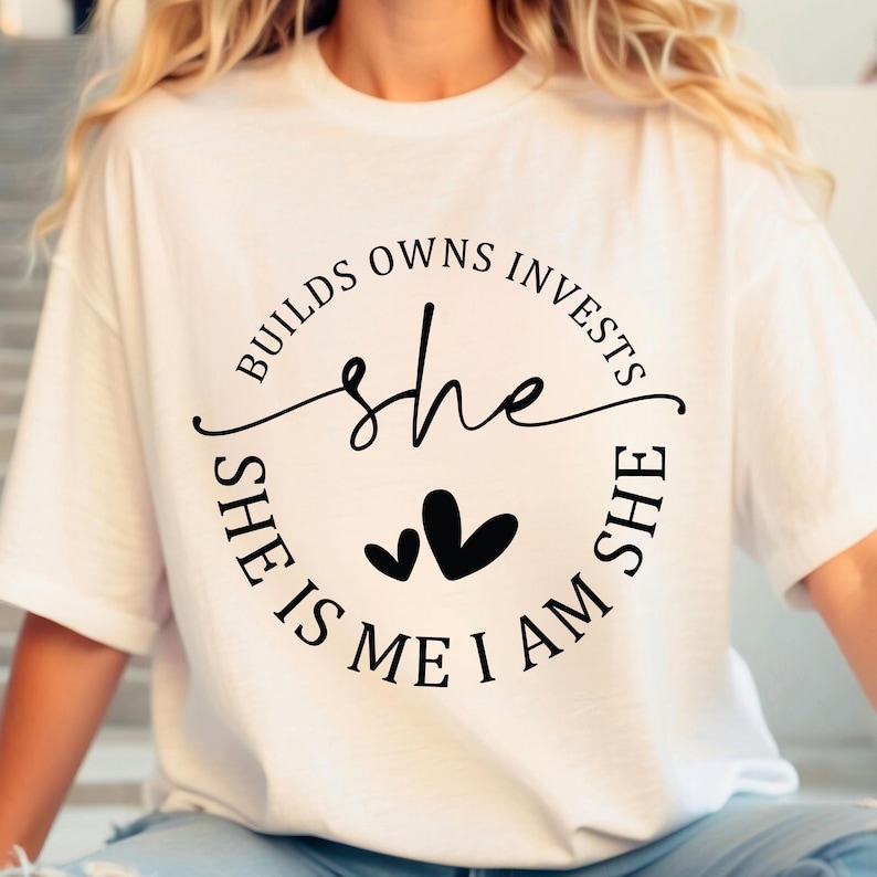 May include: White t-shirt with a black circle graphic that says "She Builds Owns Invests She Is Me I Am She" with two small hearts inside the circle.