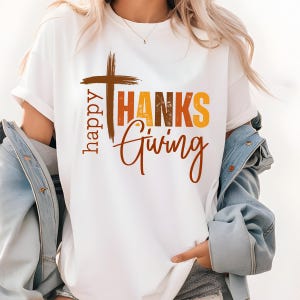 May include: White t-shirt with a graphic design that says "happy THANKS giving" with a brown cross above the word "THANKS". The text is in a vintage style with orange, yellow, and brown colors.