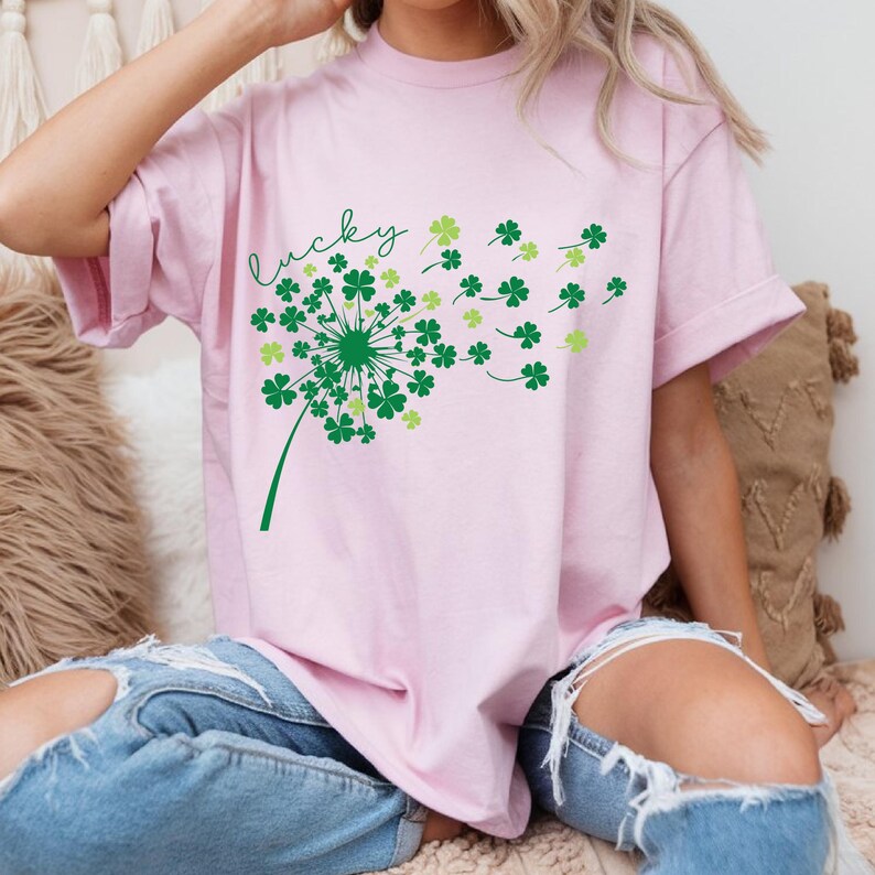 Puede incluir: Una camiseta rosa con un dise&ntilde;o de diente de le&oacute;n de tr&eacute;bol de cuatro hojas verde y la palabra "lucky" en cursiva.