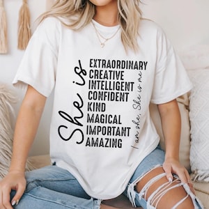 May include: White t-shirt with black text that reads "She is..." and lists positive qualities like "extraordinary", "creative", "intelligent", "confident", "kind", "magical", "important", and "amazing". The text "I am she, She is me" is on the right side of the shirt.