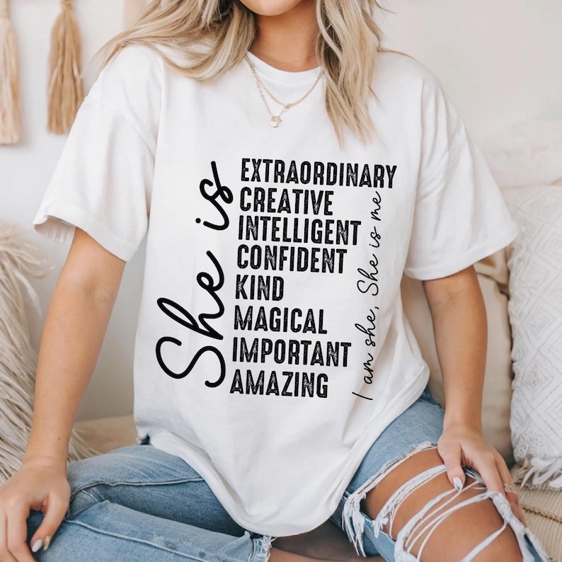 May include: White t-shirt with black text that reads "She is..." and lists positive qualities like "extraordinary", "creative", "intelligent", "confident", "kind", "magical", "important", and "amazing". The text "I am she, She is me" is on the right side of the shirt.