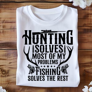 Può includere: Maglietta bianca con un design grafico nero che dice "La caccia risolve la maggior parte dei miei problemi. La pesca risolve il resto." La grafica include anche un fucile, corna di cervo e pesci.