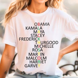 Może przedstawiać: Biała koszulka z czarnym, czerwonym, ż&oacute;łtym i zielonym wzorem. Wz&oacute;r tworzy napis "BLACK HISTORY" wykorzystując nazwiska znanych Afroamerykan&oacute;w, w tym Obamy, Kamali, Mai, Stacey, Fredericka, Thurgooda, Michelle, Rosy, Martina, Malcolma, Harriet i Garvey'ego.