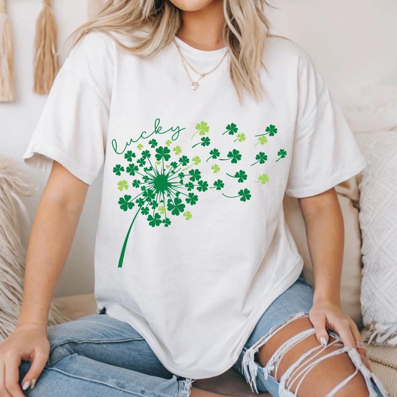 Puede incluir: Camiseta blanca con un dise&ntilde;o de diente de le&oacute;n de tr&eacute;bol verde y la palabra "lucky" escrita en cursiva.