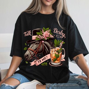 以下が含まれることがあります： 黒いTシャツにピンクの花柄、帽子をかぶった馬、そして「Talk Derby To Me!」の文字が書かれています