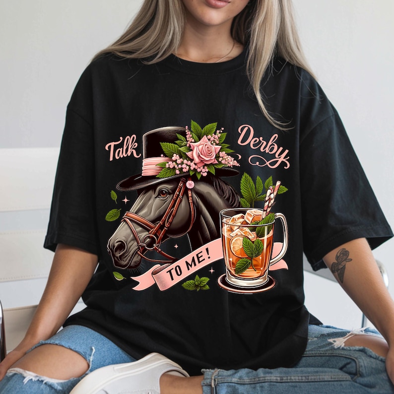 以下が含まれることがあります： 黒いTシャツにピンクの花柄、帽子をかぶった馬、そして「Talk Derby To Me!」の文字が書かれています