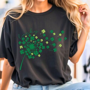 Puede incluir: Camiseta negra con un dise&ntilde;o de tr&eacute;bol verde y la palabra "lucky" escrita en cursiva. Los tr&eacute;boles est&aacute;n dispuestos en forma de diente de le&oacute;n.