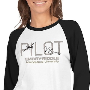 May include: A white and black baseball style shirt with a gray graphic that says "Pilot Mom" with a helicopter graphic and the text "Embry-Riddle Aeronautical University" underneath.