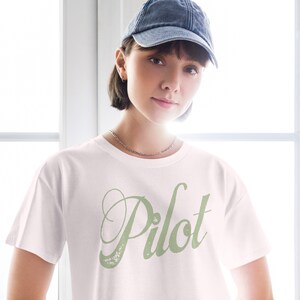 May include: A white t-shirt with a green cursive word "Pilot" printed on the front. The person wearing the shirt is also wearing a blue denim baseball cap.