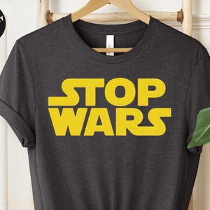 Puede incluir: Una camiseta gris oscuro jaspeada con el texto "STOP WARS" en letras amarillas. La camiseta es de cuello redondo con mangas cortas.