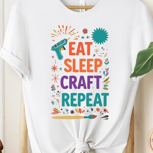 May include: White t-shirt with the text "EAT SLEEP CRAFT REPEAT" in colorful letters. The design includes a glue gun, stars, and floral elements. The shirt is displayed on a wooden hanger.