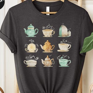 May include: A heather dark gray t-shirt with a graphic of various teapots and teacups in shades of green, blue, and gold. The graphic is centered on the shirt and features a watercolor-like style.