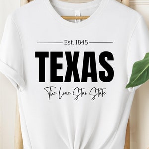 Könnte beinhalten: Weißes T-Shirt mit schwarzem Text, der "Est. 1845 Texas The Lone Star State" lautet.