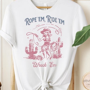 May include: White t-shirt with a vintage graphic of a cowgirl roping a calf. The graphic says "Rope 'Em, Ride 'Em, Wreck 'Em".