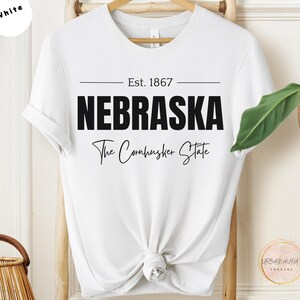 Op de afbeelding: Wit T-shirt met zwarte tekst die "Est. 1867 Nebraska The Cornhusker State" luidt