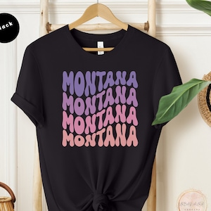 以下が含まれることがあります： 黒いTシャツ。紫からピンクのグラデーションで「MONTANA」の文字が波状に4回重ねてデザインされています。木製のハンガーにかけられています。デザインはレトロスタイルです。