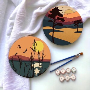 May include: Two round wooden canvases with painted sunset scenes. One canvas features a lake with tall grass and dragonflies, while the other depicts a mountain range with a single tree. Both canvases have a yellow sun and a blue sky.