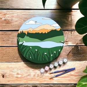 May include: A round wooden canvas with a painted landscape scene of a lake, mountains, and trees. The canvas is surrounded by six small paint pots with numbers 1 through 6.