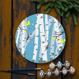 May include: A round paint-by-numbers kit featuring a winter birch forest scene with two woodpeckers. The kit includes a pre-printed canvas, paint pots, and paintbrushes. The color palette includes blue, white, black, red, and yellow.