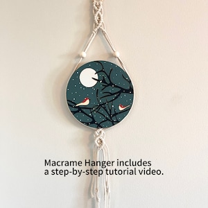 May include: A round, teal-colored wall hanging with a night scene featuring a full moon, bare tree branches, and two red birds. The artwork is suspended by a macrame hanger with wooden beads. Text on the image reads: Macrame Hanger includes a step-by-step tutorial video.
