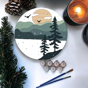 May include: A round paint-by-numbers canvas with a winter scene of two pine trees in front of a mountain range with a yellow sun and two birds flying in the sky. The canvas is surrounded by paint pots and brushes.