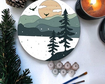 Puede incluir: Un lienzo redondo de pintura por números con una escena de invierno con dos pinos frente a una cordillera, un sol amarillo y dos pájaros volando en el cielo. El lienzo está rodeado de botes de pintura y pinceles.