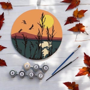 May include: A circular paint-by-number canvas featuring a sunset landscape with a yellow sun, orange sky, dark mountains, and a lake. Silhouettes of tall reeds and dragonflies are in the foreground. Includes seven numbered paint pots and two brushes.