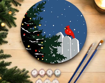 Puede incluir: Un kit de pintura por números que representa una escena de invierno con un cardenal rojo posado en una valla blanca. Un árbol de Navidad con adornos rojos y amarillos está en primer plano. El kit incluye siete botes de pintura y dos pinceles.