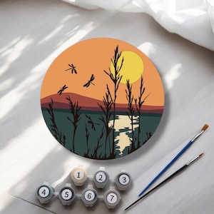 Op de afbeelding: Een ronde paint-by-numbers kit met een zonsonderganglandschap met libellen. Het kunstwerk heeft een kleurenschema van oranje en blauwgroen met een gele zon. Inclusief genummerde verfpotten (1-7) en twee penselen.