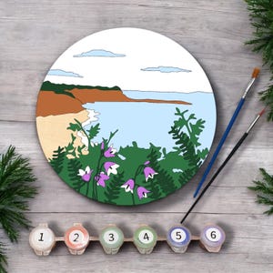 Op de afbeelding: Een rond doek voor schilderen op nummer met een kustlandschap met een strand, zee, kliffen en bloemen. Het doek is omgeven door zes nummerpotjes van 1 tot en met 6.