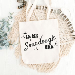 K&ouml;nnte beinhalten: Eine wei&szlig;e Stofftasche mit schwarzem Text, der "In My Sourdough Era" lautet, mit funkelnden Sternen um den Text herum.