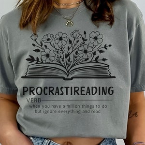 May include: A gray t-shirt with a black graphic of an open book with flowers growing out of it. The text "PROCRASTIREADING" is above the book and the text "VERB" is above the text "PROCRASTIREADING". The text below the book reads "when you have a million things to do but ignore everything and read."