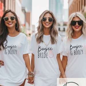 Może przedstawiać: Trzy przyjaciółki w białych koszulkach z nadrukiem "Boujee & Boozy" i "Boujee Bride". Koszulka panny młodej ma grafikę różowego pierścionka. Noszą brązowe kozaki za kolano i uśmiechają się.