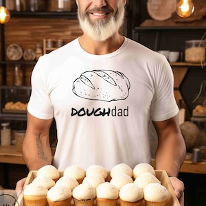 May include: A white t-shirt with a black and white illustration of a loaf of bread and the text "DOUGHdad". A person is holding a tray of cupcakes with white frosting.