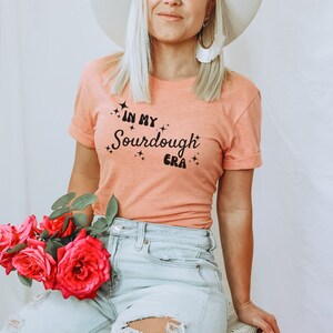 K&ouml;nnte beinhalten: Eine Frau tr&auml;gt ein pfirsichfarbenes T-Shirt mit dem Text "In My Sourdough Era" in schwarzen Buchstaben. Sie tr&auml;gt einen wei&szlig;en Hut und eine blaue Jeans.